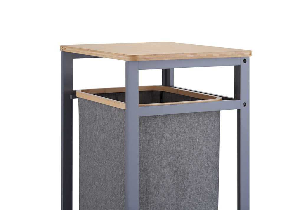 A close-up shot highlights the upper portion of a laundry hamper unit, focusing on its light wooden tabletop and dark gray metal frame. Directly beneath the top shelf, a gray fabric hamper bag with a wooden-trimmed rim rests suspended inside the structure.