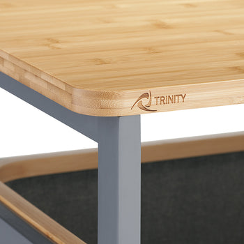 A close-up photograph highlights the rounded corner of a light bamboo tabletop mounted on a dark gray metal frame. The front edge of the wooden surface is subtly engraved with the brand logo "TRINITY".