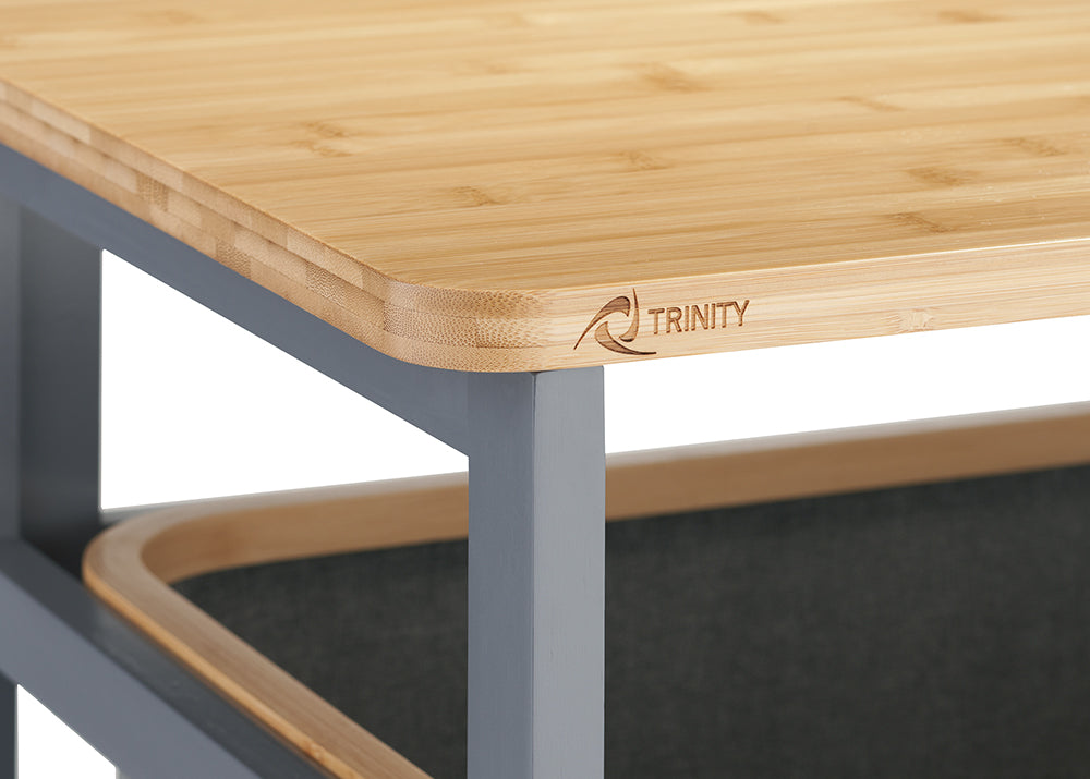 A close-up photograph highlights the rounded corner of a light bamboo tabletop mounted on a dark gray metal frame. The front edge of the wooden surface is subtly engraved with the brand logo "TRINITY".