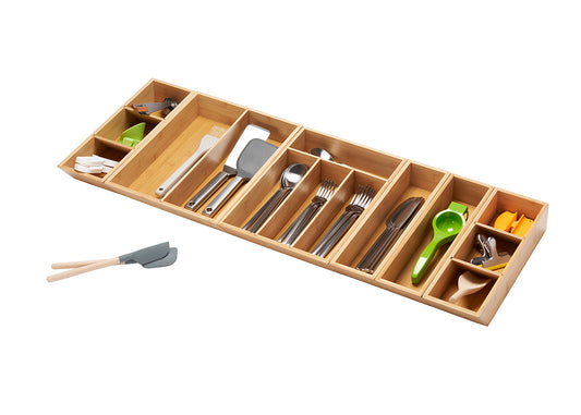A top-down, angled view shows a light brown bamboo drawer organizer fully extended and filled with various kitchen utensils. Outside the organizer, to the left, are two small gray spatulas with light brown wooden handles.