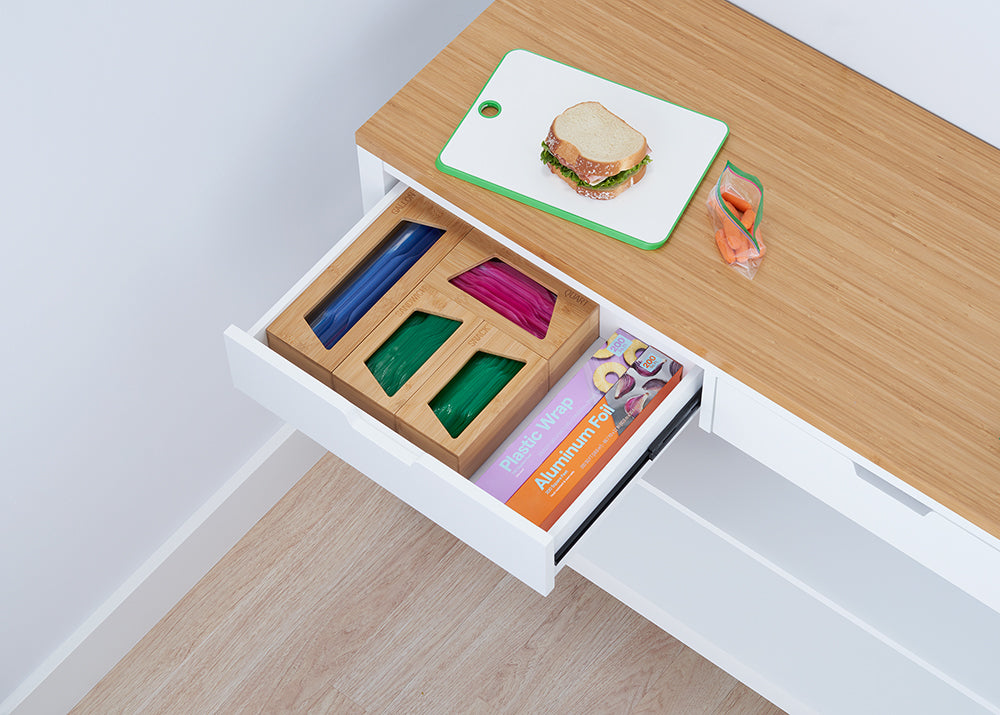 A drawer beneath a light wood countertop is pulled open, revealing a wooden organizer holding colorful plastic bags and a box of aluminum foil and plastic wrap. On the countertop sits a sandwich on a cutting board and a plastic bag of carrots, suggesting a snack preparation area.