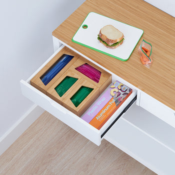 A drawer beneath a light wood countertop is pulled open, revealing a wooden organizer holding colorful plastic bags and a box of aluminum foil and plastic wrap. On the countertop sits a sandwich on a cutting board and a plastic bag of carrots, suggesting a snack preparation area.