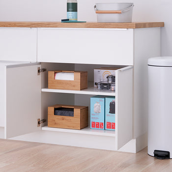 A slightly ajar white cabinet with two shelves reveals its contents: two small wooden boxes and two blue and pink cardboard boxes. To the right of the cabinet is a tall, cylindrical white trash can with a black foot pedal.