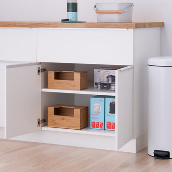 A slightly ajar white cabinet under a light wood countertop reveals two shelves holding various items, including wooden boxes and packaged goods. A white trash can stands to the right of the cabinet, and the scene is set against a plain white wall.