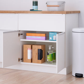 A partially open white cabinet under a light wood countertop reveals a shelf with various household items. To the right of the cabinet is a tall, white trash can with a black foot pedal.