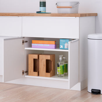 A slightly ajar white cabinet with a light wood countertop reveals two shelves holding various household items, including cleaning supplies, boxes, and wooden organizers. To the right of the cabinet stands a tall, white trash can with a black foot pedal.