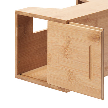 A close-up shot shows a rectangular bamboo trash bag dispenser with a partially open sliding door. The light-colored bamboo has a visible wood grain, and the dispenser has a cutout on the top and a dispensing slot on the side.