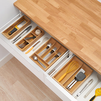 A top-down view of two kitchen drawers pulled open to reveal a tidy organization of utensils. In the drawers are wooden dividers separating various kitchen tools, including silverware, whisks, and graters.