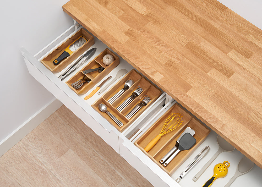 A top-down view of two kitchen drawers pulled open to reveal a tidy organization of utensils. In the drawers are wooden dividers separating various kitchen tools, including silverware, whisks, and graters.