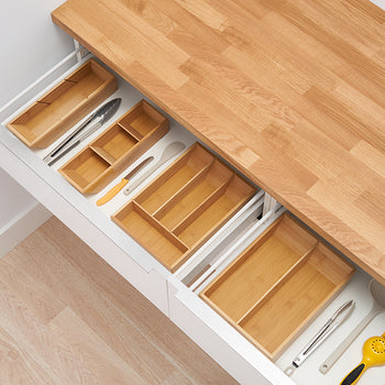 A top-down view of two kitchen drawers with various wooden organizers. The top drawer is partially open, revealing a utensil organizer, while the bottom drawer is fully extended, showing larger, empty wooden trays and some kitchen tools.