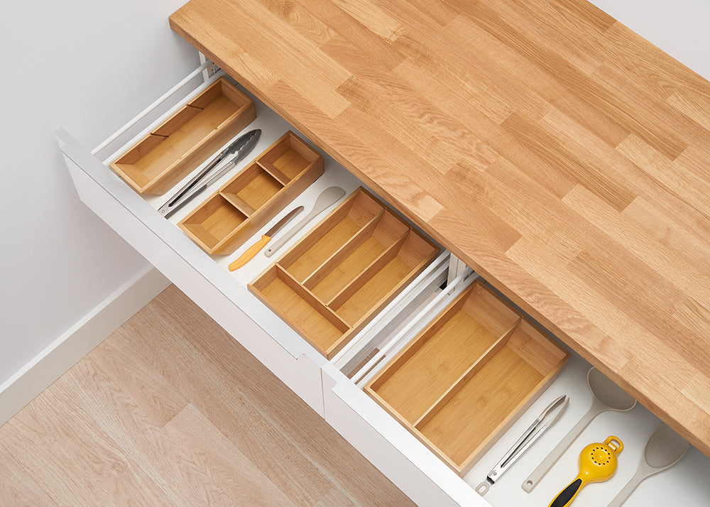 A top-down view of two kitchen drawers with various wooden organizers. The top drawer is partially open, revealing a utensil organizer, while the bottom drawer is fully extended, showing larger, empty wooden trays and some kitchen tools.