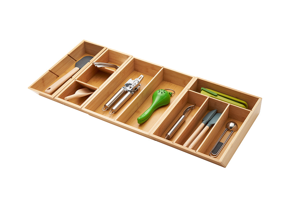A high-angle shot of a rectangular, adjustable bamboo drawer organizer filled with various kitchen utensils. The organizer has five main compartments, with an additional smaller compartment on the far left.