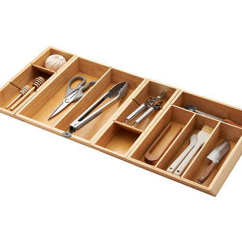 A close-up, high-angle shot of a bamboo drawer organizer with various kitchen tools neatly arranged inside. The organizer is filled with items like a honey dipper, scissors, tongs, a can opener, basting brushes, and a knife.