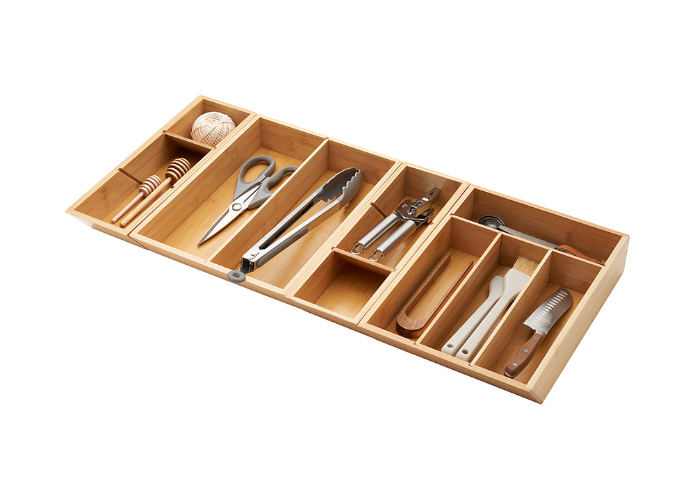 A close-up, high-angle shot of a bamboo drawer organizer with various kitchen tools neatly arranged inside. The organizer is filled with items like a honey dipper, scissors, tongs, a can opener, basting brushes, and a knife.