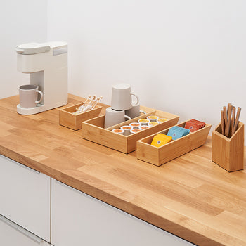 A clean, organized coffee station is set up on a wooden countertop. It features a single-serve coffee maker and several bamboo organizers holding coffee pods, cups, stirrers, and other coffee-related items.