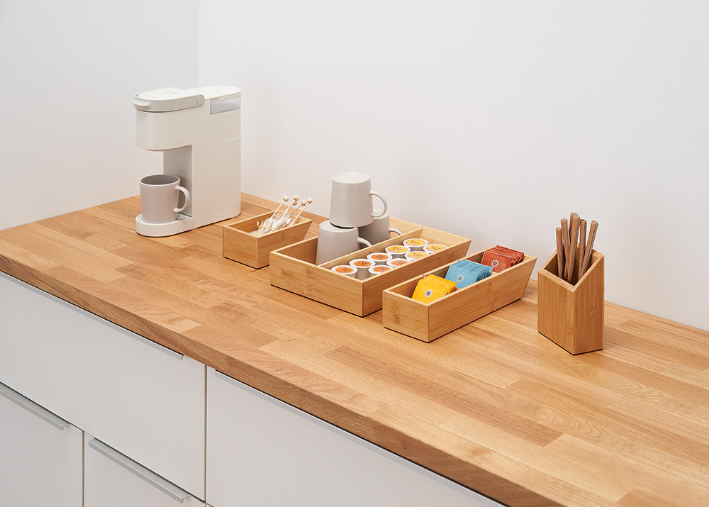 A clean, organized coffee station is set up on a wooden countertop. It features a single-serve coffee maker and several bamboo organizers holding coffee pods, cups, stirrers, and other coffee-related items.