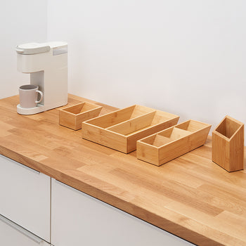 A white coffee maker with a gray mug sits on a wooden countertop next to a set of four light-colored bamboo storage containers. The containers vary in size and shape, with one rectangular, one long and divided into two sections, and one smaller and square.