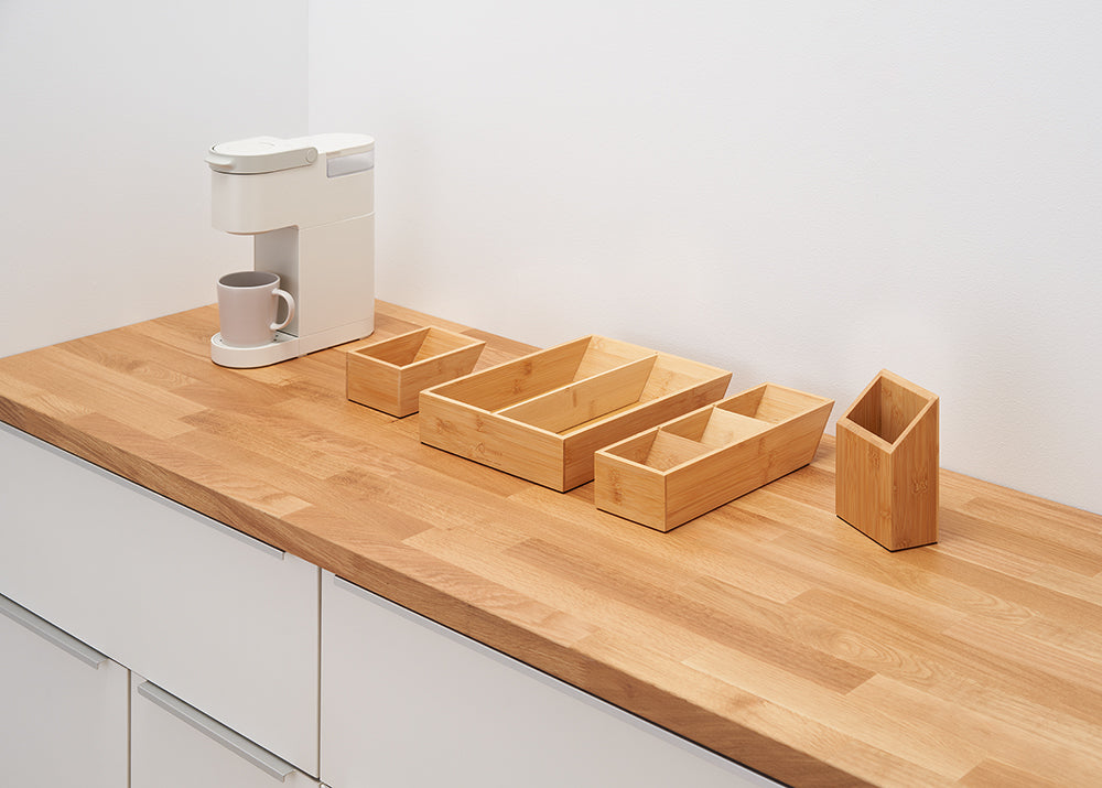 A white coffee maker with a gray mug sits on a wooden countertop next to a set of four light-colored bamboo storage containers. The containers vary in size and shape, with one rectangular, one long and divided into two sections, and one smaller and square.