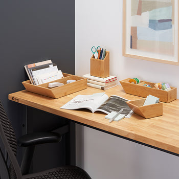 A desk is organized with several bamboo organizers holding office supplies and paperwork. The desk is made of light wood with a dark gray and white wall behind it, and a framed abstract picture hangs on the white wall.