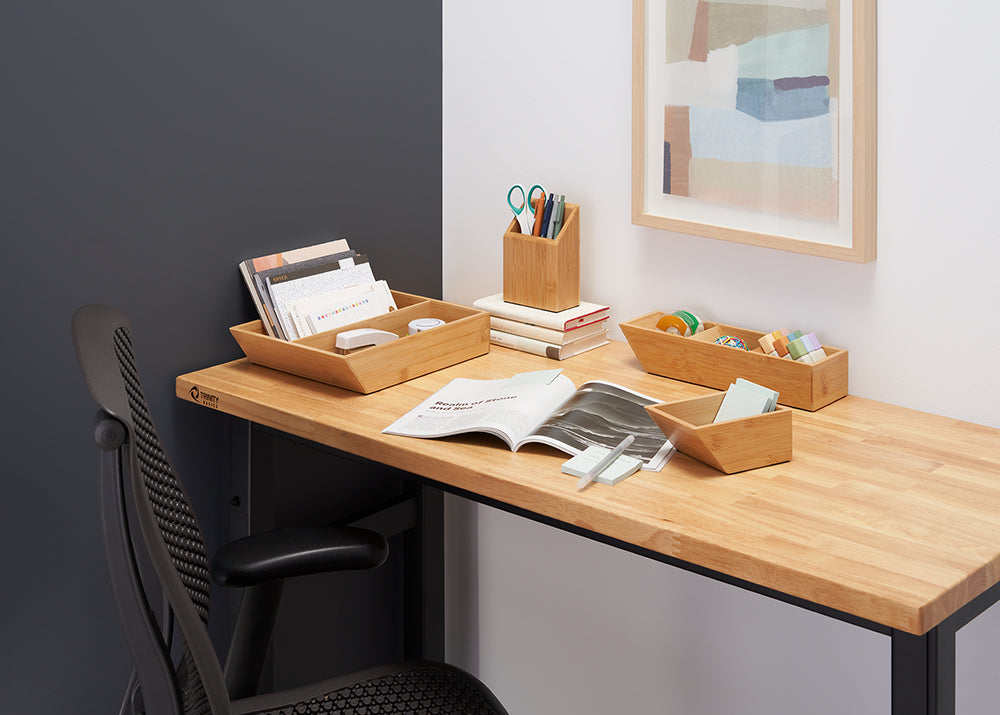 A desk is organized with several bamboo organizers holding office supplies and paperwork. The desk is made of light wood with a dark gray and white wall behind it, and a framed abstract picture hangs on the white wall.