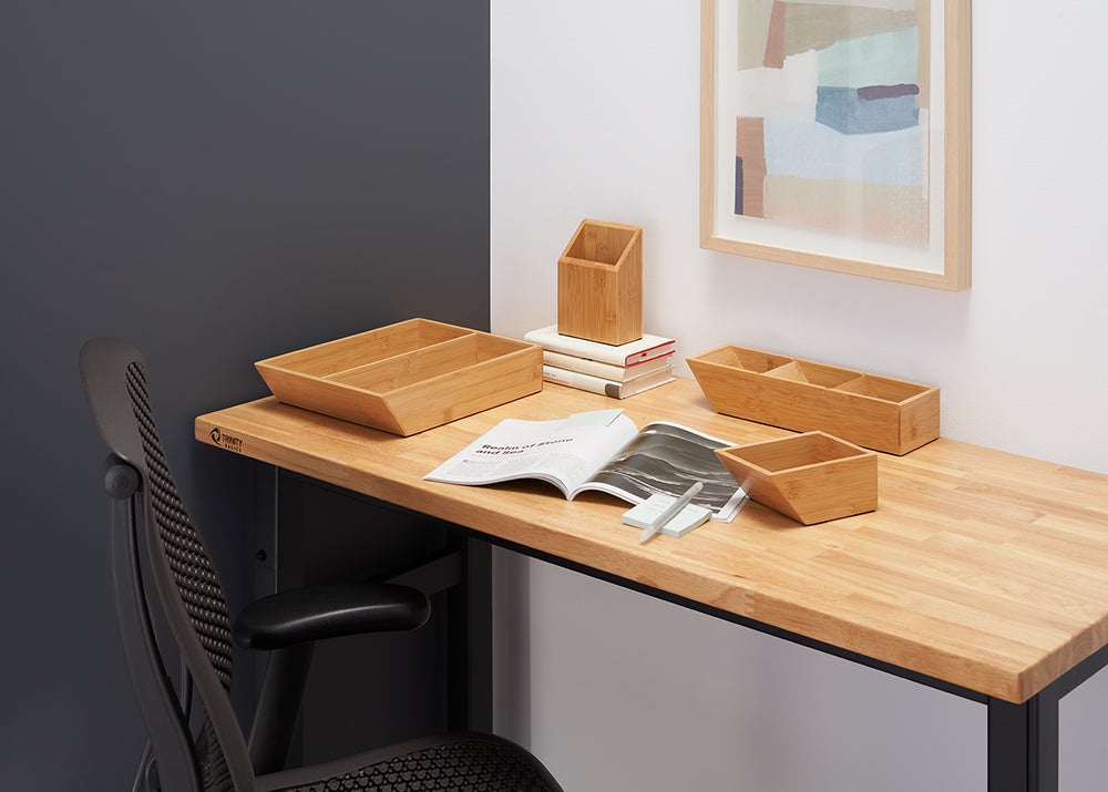 A minimalist home office desk is shown with several bamboo organizers, including a shallow divided tray and a deeper upright container. The wooden desktop also holds a few books, an open magazine, and a small stack of sticky notes.