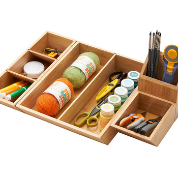 A high-angle shot shows a wooden craft organizer with multiple compartments filled with various crafting supplies. The organizer contains yarn, markers, scissors, paintbrushes, paints, and other small tools.