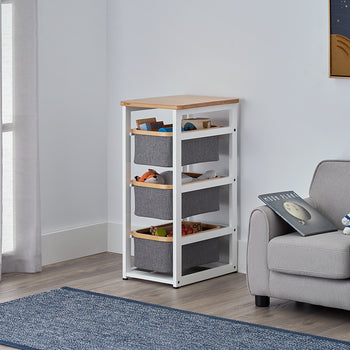 A white-framed storage unit with three grey fabric bins and a wood-grain top sits in the corner of a child's room. Beside it is a grey armchair holding a book and an astronaut teddy bear, all set against a neutral wall with a space-themed poster.