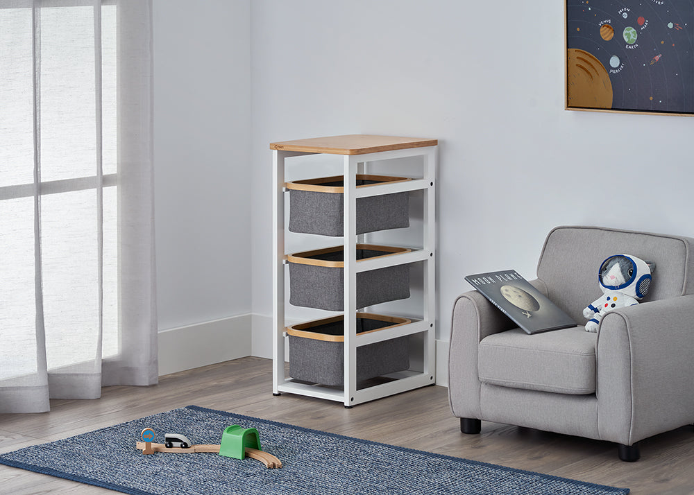 A modern white storage rack with a wooden top and three grey fabric bins stands in the corner of a child's space-themed room. The setting includes a grey armchair with an astronaut plush, a blue rug with a toy train, and a solar system poster on the wall.