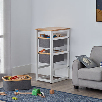 A white wooden storage unit with a light wood top and three grey fabric bins stands in a bright, modern nursery. The surrounding room features a matching grey armchair with an astronaut plush toy, a blue rug with wooden train tracks, and a solar system poster on the wall.