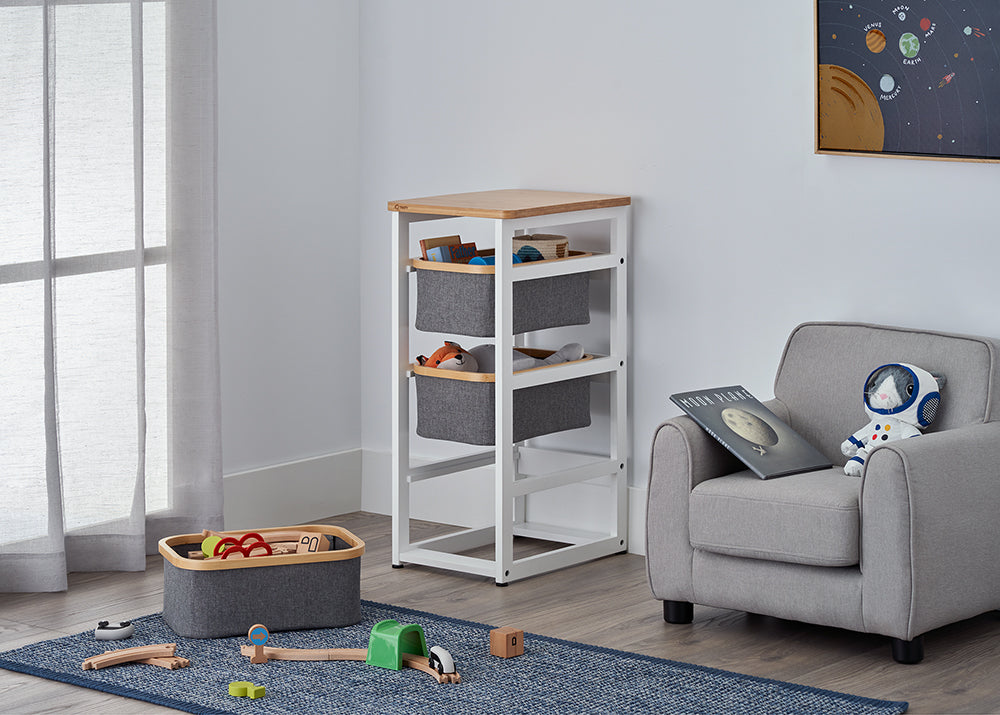 A white wooden storage unit with a light wood top and three grey fabric bins stands in a bright, modern nursery. The surrounding room features a matching grey armchair with an astronaut plush toy, a blue rug with wooden train tracks, and a solar system poster on the wall.