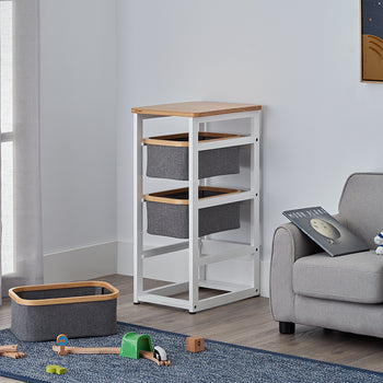 A white three-tier storage rack with a wood top and grey bins sits in the corner of a child's room next to a small grey armchair. On a blue rug in the foreground, a matching grey bin sits near a wooden toy train set and a green tunnel.