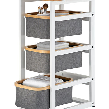 A multi-tiered storage organizer features a white metal frame with a light wood tabletop and three pull-out grey fabric bins. Each bin is accented with a wooden rim and holds various bathroom essentials, including makeup brushes, towels, and toiletries.