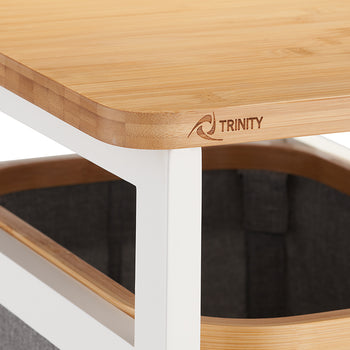 A close-up shot focuses on the corner of a light-colored bamboo tabletop featuring a small, engraved "TRINITY" logo. Beneath the wooden surface, a dark grey fabric storage bin with a matching bamboo rim is partially visible within a white metal frame.