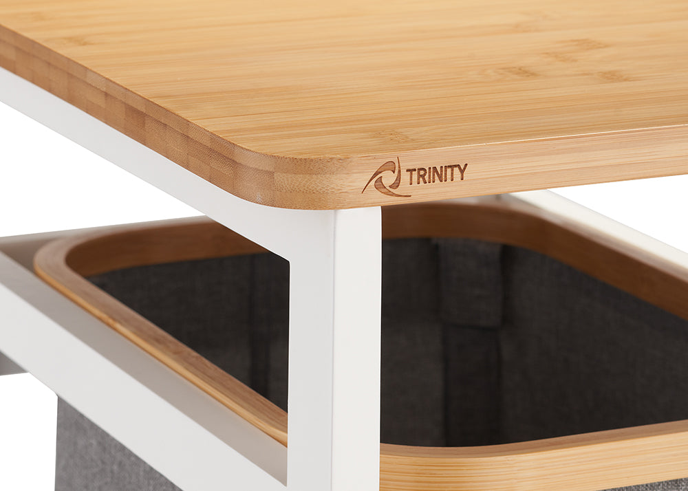 A close-up shot focuses on the corner of a light-colored bamboo tabletop featuring a small, engraved "TRINITY" logo. Beneath the wooden surface, a dark grey fabric storage bin with a matching bamboo rim is partially visible within a white metal frame.