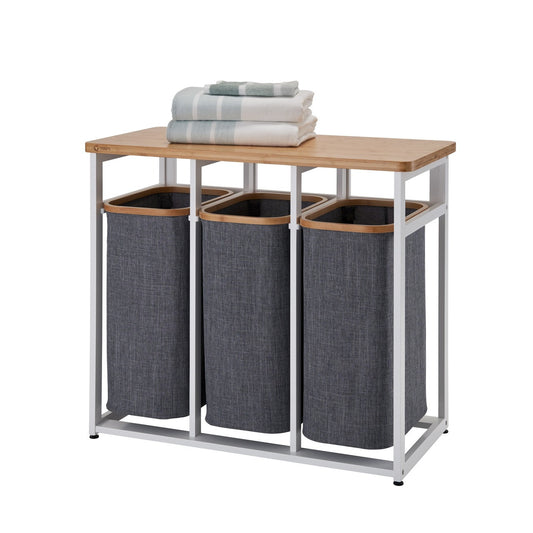 A three-compartment laundry sorter with a sturdy black metal frame, dark gray fabric hampers with wooden rims, and a light-colored wooden top surface. The top holds a detergent bottle and a glass jar with wooden clothespins.