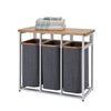 A three-compartment laundry sorter with a sturdy black metal frame, dark gray fabric hampers with wooden rims, and a light-colored wooden top surface. The top holds a detergent bottle and a glass jar with wooden clothespins.