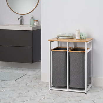 A close-up shot of a modern laundry hamper stand with two grey cloth baskets and a light wood top, set against a white wall with a minimalist sink and mirror in the background. The stand is holding folded towels, a loofah, and a bottle of soap, and it is placed on a light hexagonal-tiled floor.