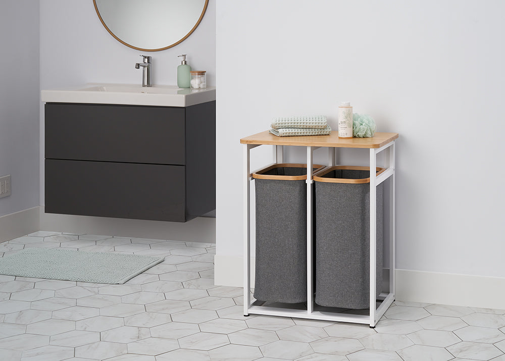 A close-up shot of a modern laundry hamper stand with two grey cloth baskets and a light wood top, set against a white wall with a minimalist sink and mirror in the background. The stand is holding folded towels, a loofah, and a bottle of soap, and it is placed on a light hexagonal-tiled floor.
