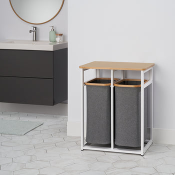 
A two-compartment laundry hamper with a natural wood top sits in a minimalist bathroom. The hamper has a white metal frame and two removable gray fabric bags.