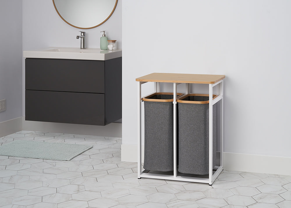 
A two-compartment laundry hamper with a natural wood top sits in a minimalist bathroom. The hamper has a white metal frame and two removable gray fabric bags.