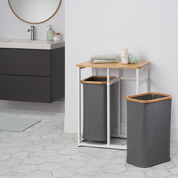 A laundry hamper set sits on a tiled floor in a modern bathroom. The set consists of a rectangular hamper and a second hamper nested within a white metal stand with a light wood top.