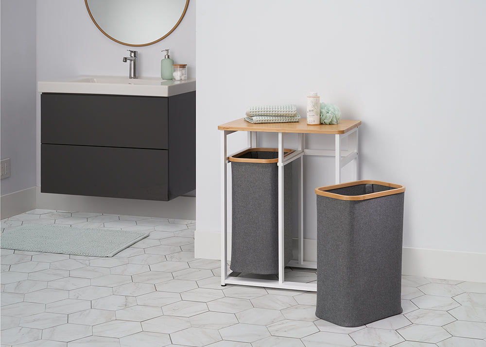 A laundry hamper set sits on a tiled floor in a modern bathroom. The set consists of a rectangular hamper and a second hamper nested within a white metal stand with a light wood top.