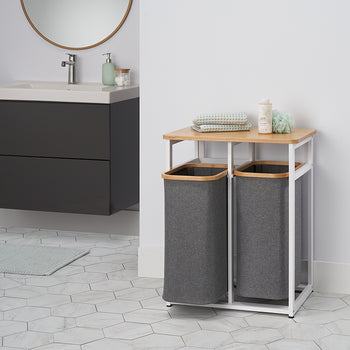 A laundry hamper with two grey fabric baskets sits on a white metal frame with a light wood top. In the background, there is a floating dark grey vanity with a white sink and a round mirror on a white tiled floor.