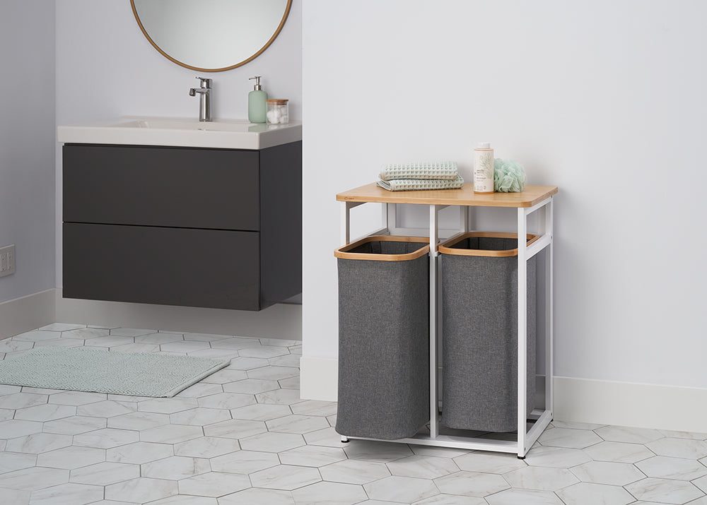 A laundry hamper with two grey fabric baskets sits on a white metal frame with a light wood top. In the background, there is a floating dark grey vanity with a white sink and a round mirror on a white tiled floor.