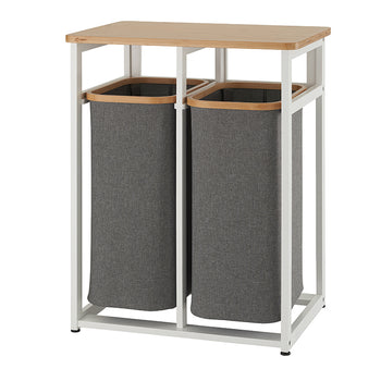 A tall, narrow laundry hamper with two separate gray fabric baskets held within a white metal frame. The top of the frame is a light-colored wood surface that can be used for folding clothes or other storage.