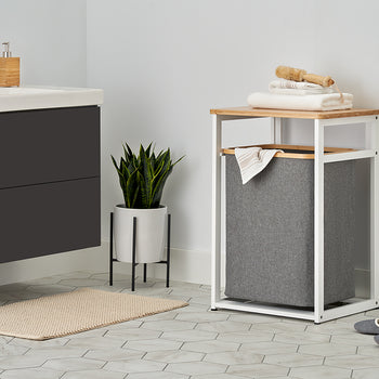 A white metal and light wood laundry hamper stand with a dark gray fabric bin sits in a modern bathroom, topped with folded white towels and a wooden brush. The surrounding bright space features light hexagonal floor tiles, a potted snake plant on a stand, a floating vanity, and a beige bath mat.