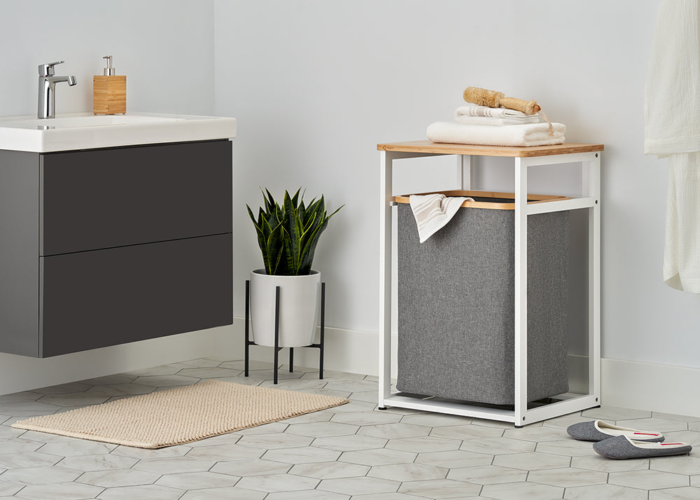 A white metal and light wood laundry hamper stand with a dark gray fabric bin sits in a modern bathroom, topped with folded white towels and a wooden brush. The surrounding bright space features light hexagonal floor tiles, a potted snake plant on a stand, a floating vanity, and a beige bath mat.