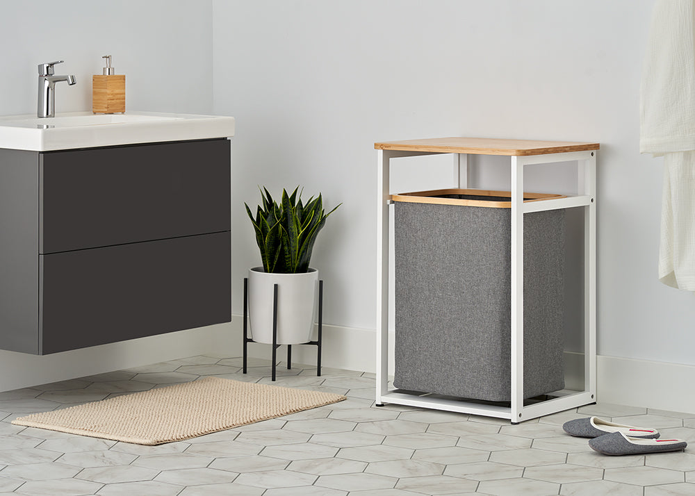 A white metal and light wood laundry hamper with a dark gray fabric bin is styled empty inside a bright, modern bathroom. The clean setup includes hexagonal tile flooring, a potted snake plant, a floating gray vanity, and a beige bath mat.