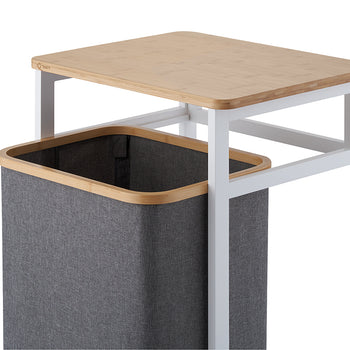 A close-up view demonstrates the dark grey fabric laundry bin sliding out from beneath the white metal frame and light wood tabletop. The high-angle shot highlights the smooth bamboo rim of the basket and the clean structure of the hamper stand against a white background.