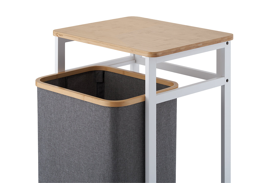 A close-up view demonstrates the dark grey fabric laundry bin sliding out from beneath the white metal frame and light wood tabletop. The high-angle shot highlights the smooth bamboo rim of the basket and the clean structure of the hamper stand against a white background.